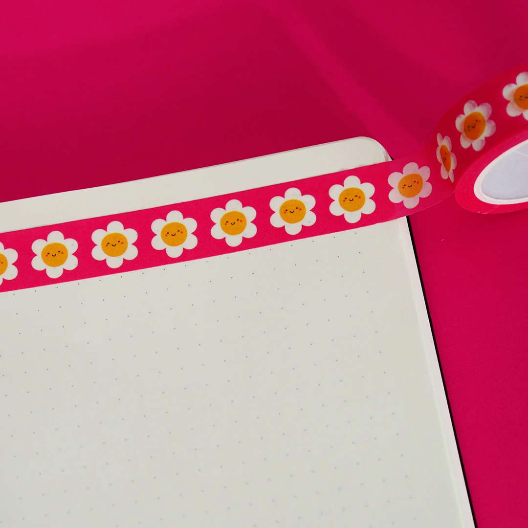 A strip of cute red washi tape daisies with smiley faces stuck onto a sheet of dotted paper on a red background