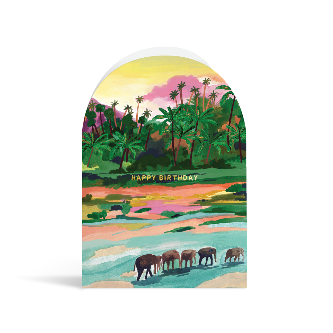 A bright, arch-shaped card painted in vibrant turquoises, greens, pinks and peach tones. The scene depicts a tropical palm forest with a group of elephants in the foreground, and gold foiled text reading Happy Birthday