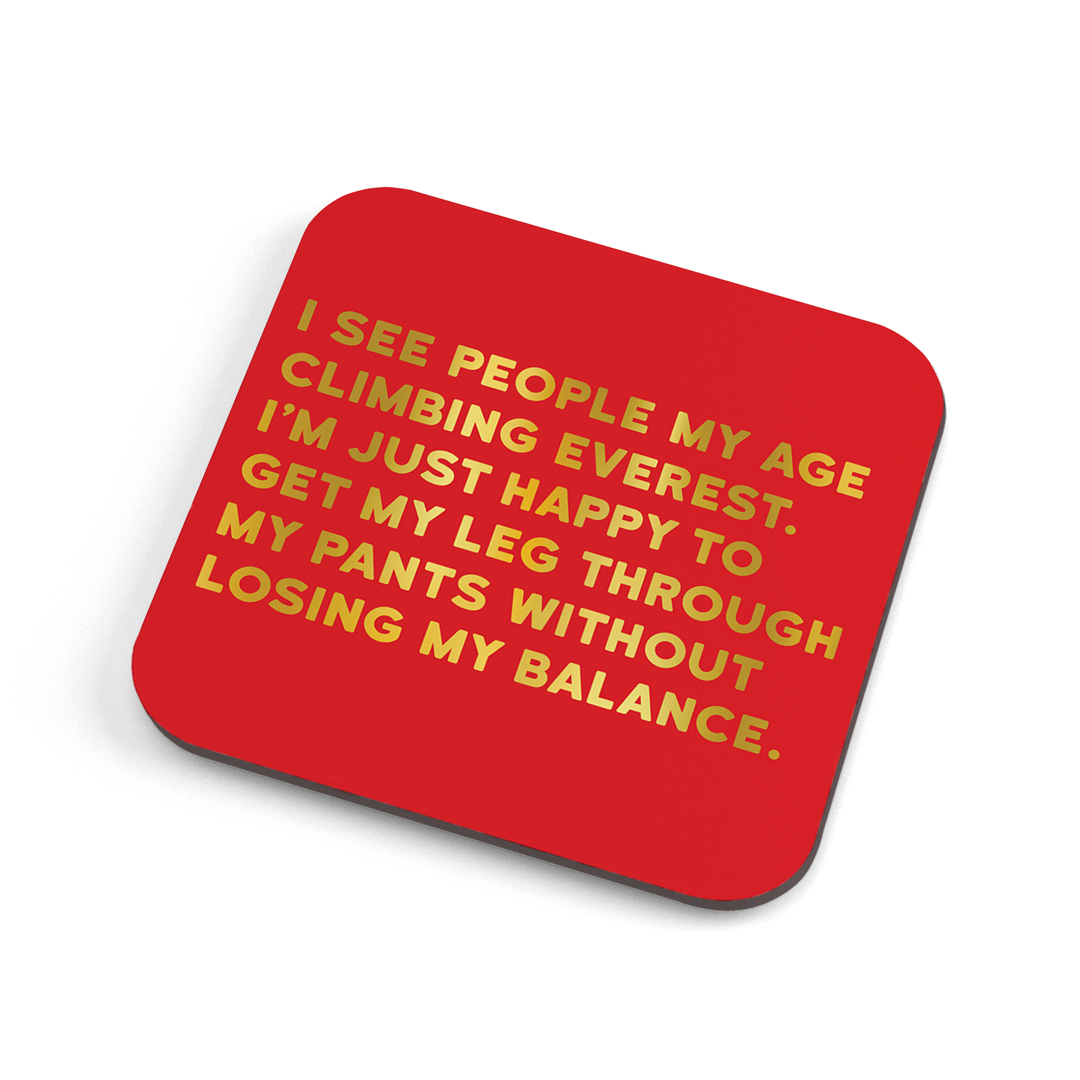 A funny red coaster with gold foiled text saying: "I see people my age climbing everest. I'm just happy to get my leg through my pants without losing my balance."