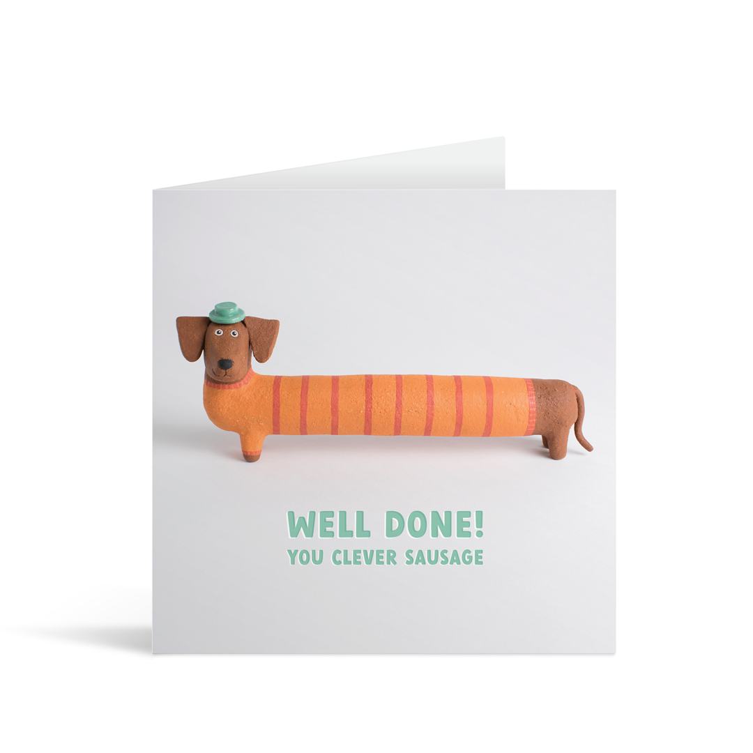 A grey square card featuring a pottery sausage dog in a cute orange and red stripey jumper and little dapper green hat. He is looking directly at the camera and standing above words that say "Well done! You clever sausage"