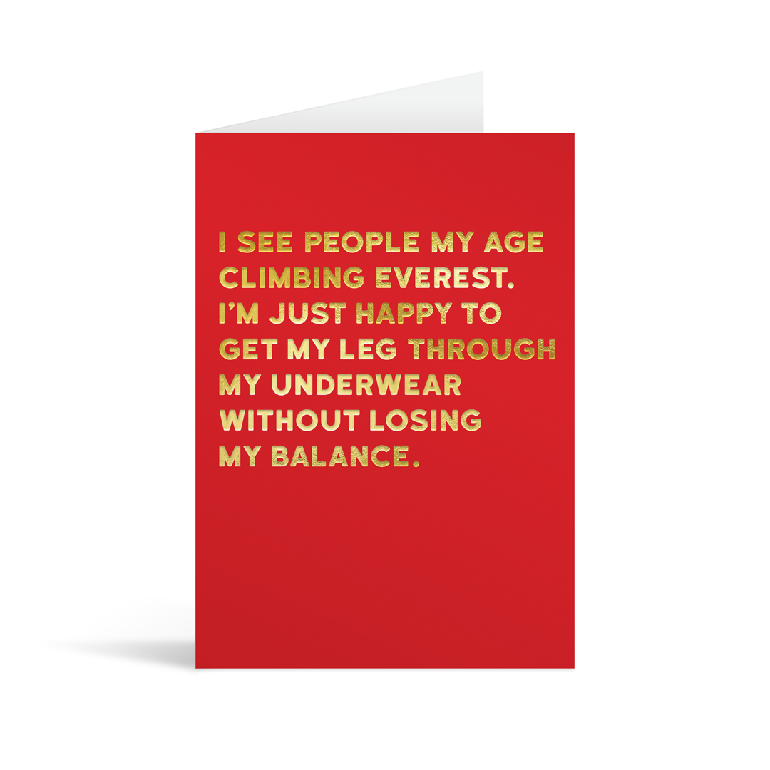 Bright red rectangular card with gold foiled text saying: " I see people my age climbing everest. I'm just happy to get my leg through my underwear without losing my balance."