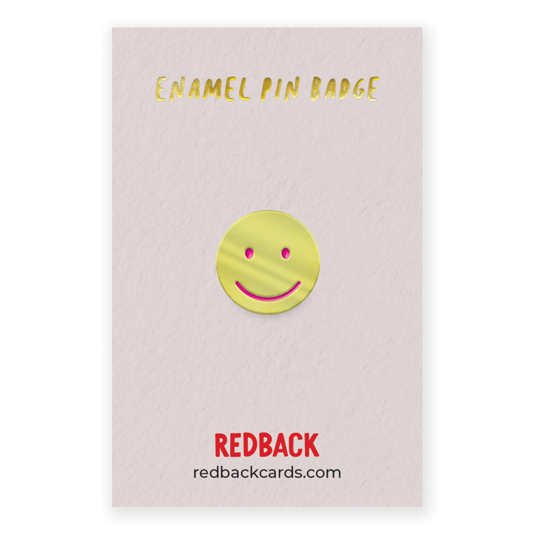 A cute gold smiley face enamel pin with pink detailing for the face. Mounted on card.