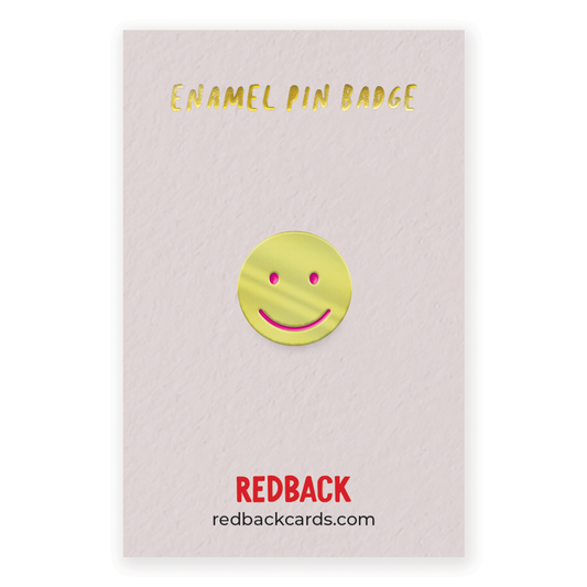 A cute gold smiley face enamel pin with pink detailing for the face. Mounted on card.