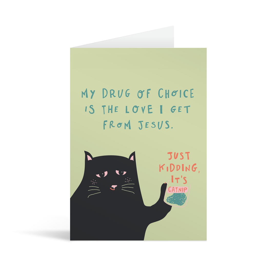 Pale sage green rectanngular card, with a quirky illustration of a cat and the handwritten text "My drug of choice is the love I get from Jesus. Just kidding, it's catnip". Brown envelope in the background.