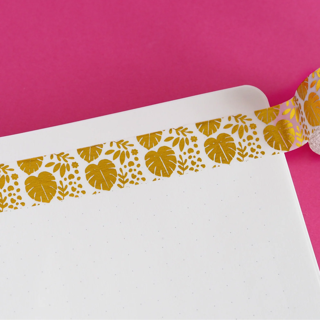 A strip of cute clear washi tape with gold foiled leaves and small flowers stuck onto a sheet of dotted paper on a red background