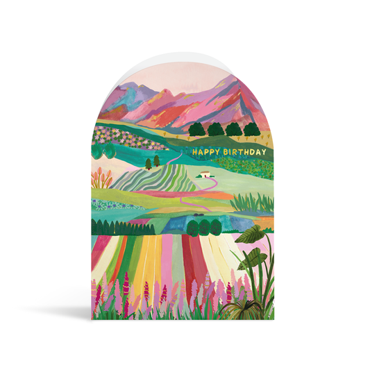 A bright, arch-shaped card painted in vibrant turquoises, greens, pinks and peach tones. The scene depicts rolling colourful fields leading away to mountains in the distance, and gold foiled text reading Happy Birthday