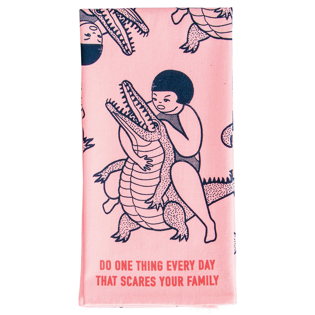 A pink tea towel or dish towel featuring a stylised image of a woman wrestling an alligator above red text saying "Do one thing every day that scares your family"