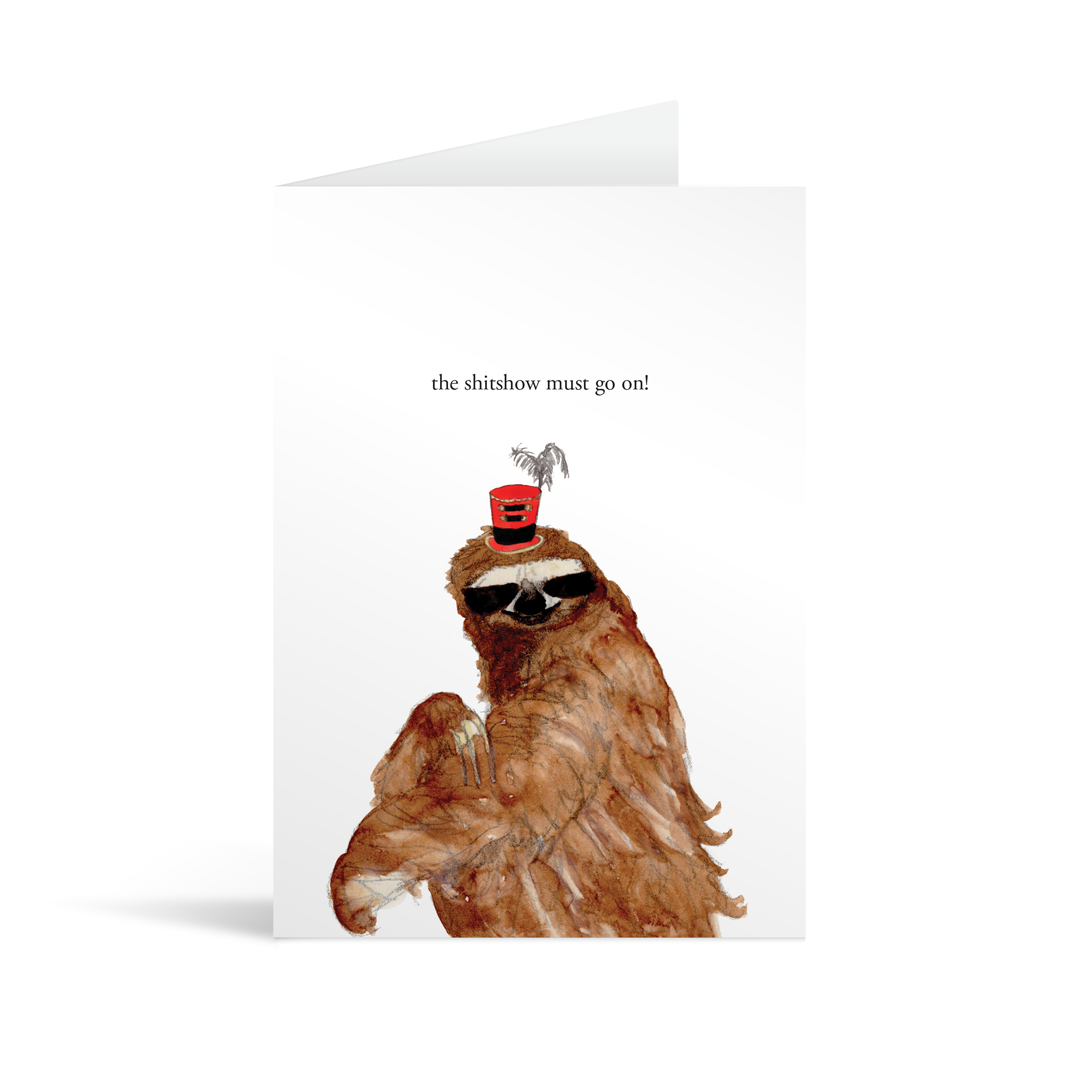 A white rectangular card with a sloth in a tophat and funny text saying "'The shitshow must go on!" , with a kraft brown envelope.