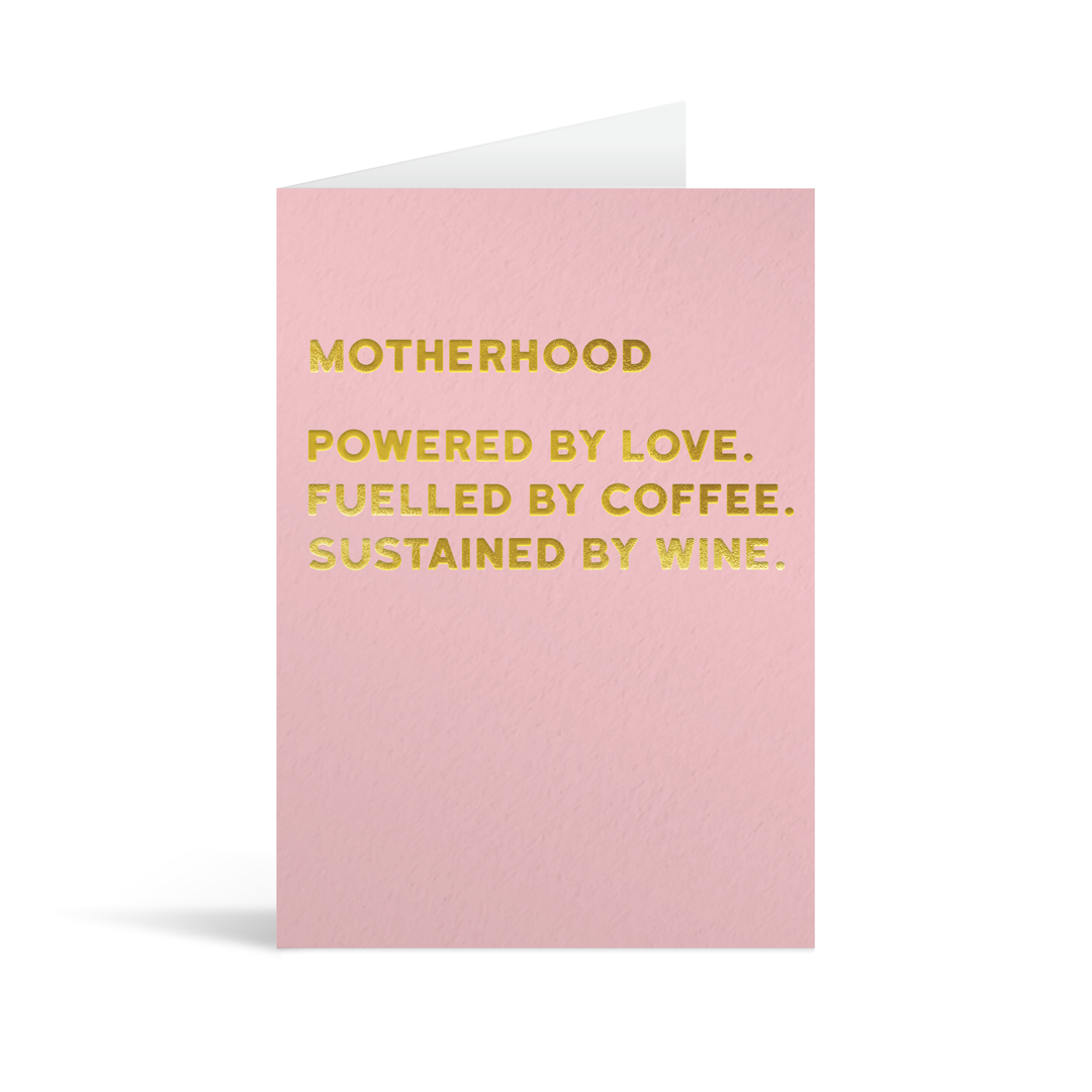 Pink rectangular card with funny gold foiled text saying "Motherhood powered by love. Fuelled by coffee. Sustained by wine."