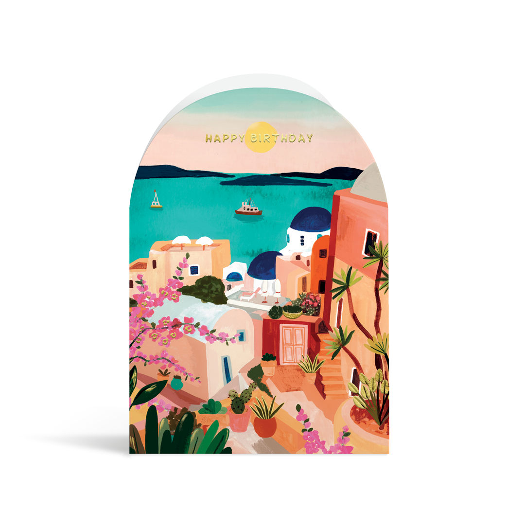 A bright, arch-shaped card painted in vibrant turquoises, pinks and peach tones. The scene depicts a typical Santorini scene with white buildings and blue roofs, and gold foiled text reading Happy Birthday