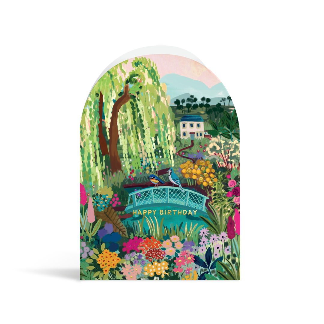 A bright, arch-shaped card painted in vibrant turquoises, greens and pink tones. The scene depicts a house overlooking a luscious garden with a bridge over a pond, and gold foiled text reading Happy Birthday