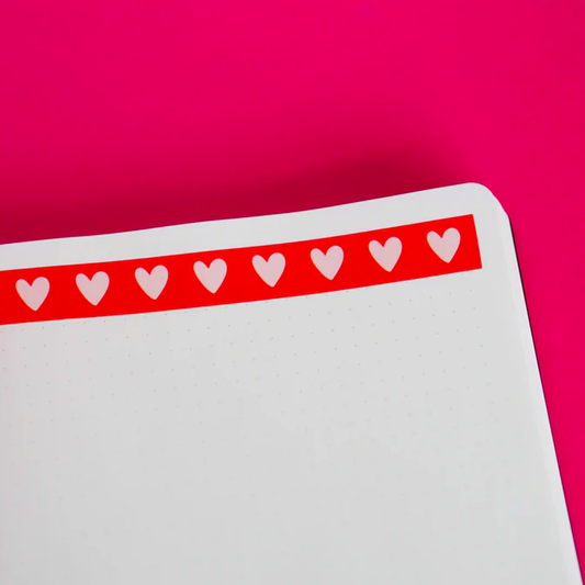 A strip of cute red washi tape with pink hearts stuck onto a sheet of dotted paper on a red background