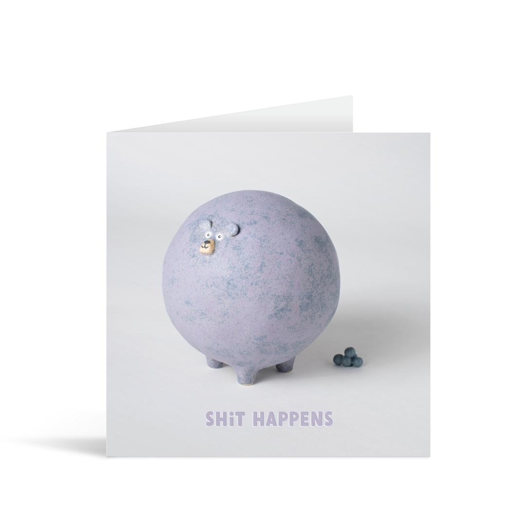 A grey square card featuring a funny pottery figure of a bloated purple bear full of character standing next to a pile of poo above the words "shit happens"